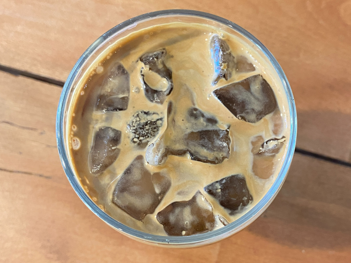 A fresh glass of iced coffee and oat milk sits on a wooden table. 