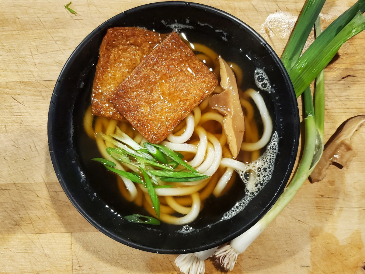 A black bowl filled with fresh udon is laying on a wooden table. The bowl is filled with fresh pressed bean tofu and udon noodles. Fresh spring onions and mushrooms are sitting on the table beside the bowl