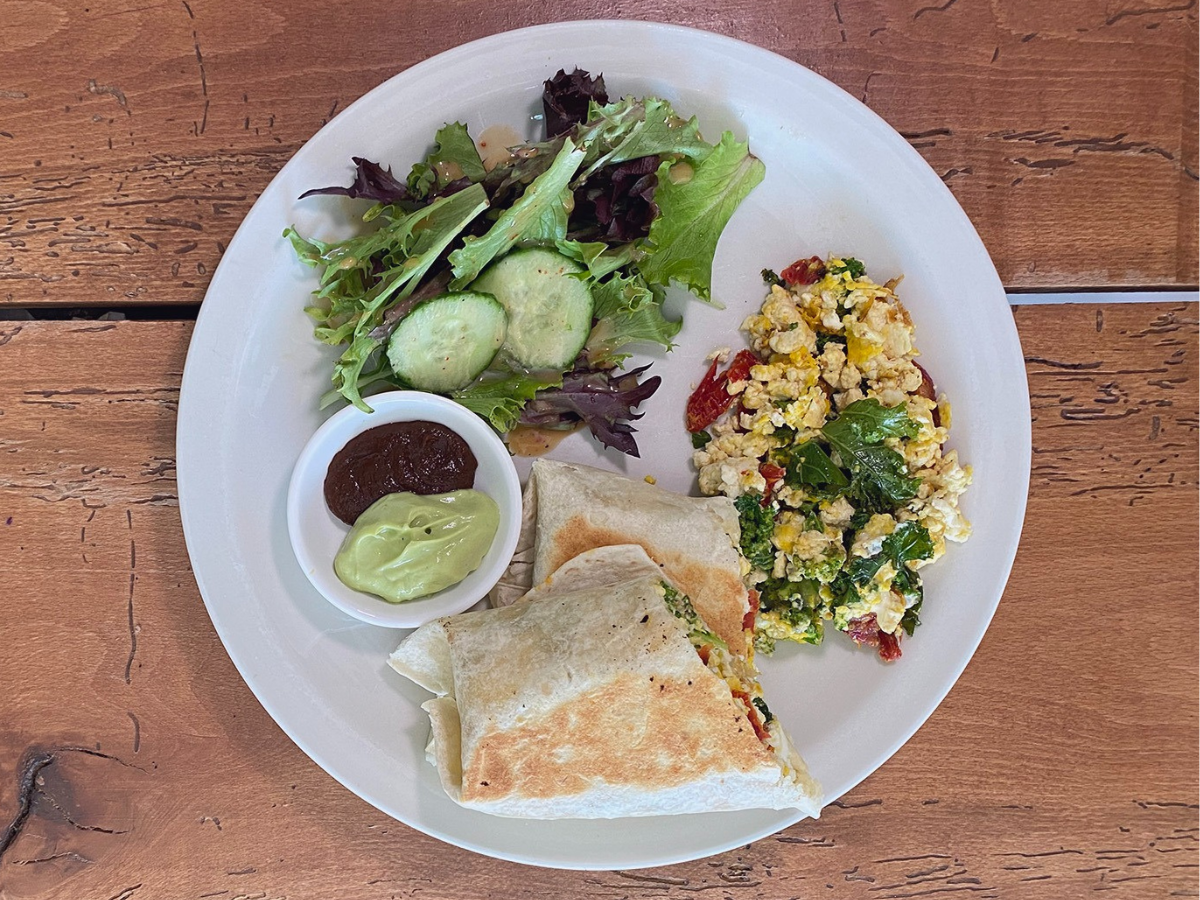 A white plate on a wooden table is filled with a crispy breakfast burrito, scrambled eggs with veggies, a side salad with sliced cucumbers, and a little finger bowl filled with guacamole and hot sauce. 