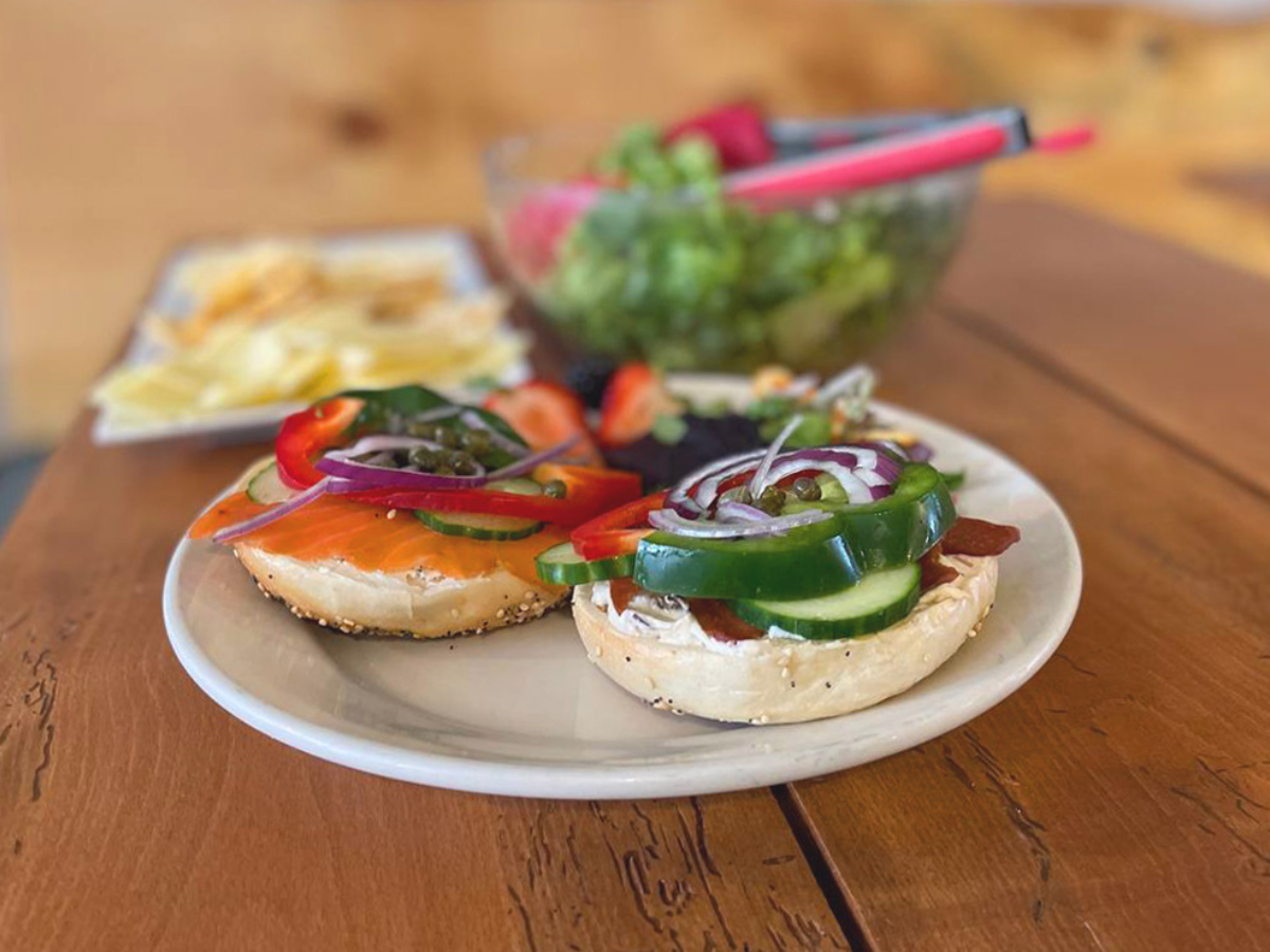 A bagel on a white plate, filled with fresh veggies and lox resting on a wooden table.